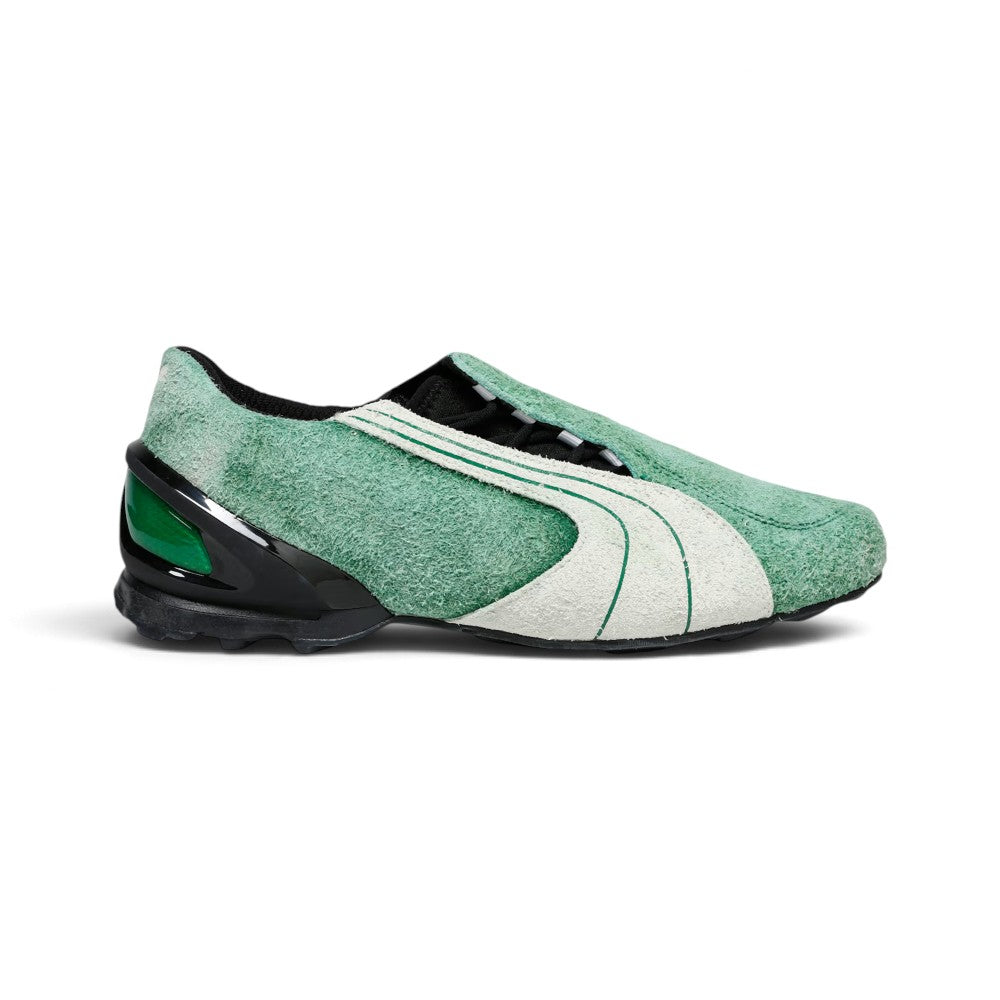 Displays the lateral profile showing the mossy-green hairy cow suede upper, double-piped white suede Formstrip, black-cased external heel counter with emerald green insert windows, concealed asymmetric lace shroud, and low-slung black turf-cleat rubber sole. Main image