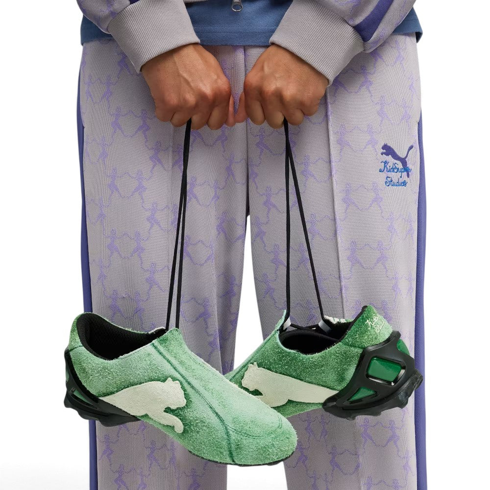 Dynamic lifestyle angle featuring the shoes suspended by their long lacing cords in front of lavender co-branded tracksuit bottoms, highlighting the asymmetric silhouette, oversized white suede medial leaping Cat emblem, and aggressive heel cage.