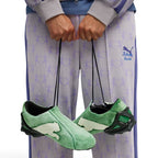 Dynamic lifestyle angle featuring the shoes suspended by their long lacing cords in front of lavender co-branded tracksuit bottoms, highlighting the asymmetric silhouette, oversized white suede medial leaping Cat emblem, and aggressive heel cage.