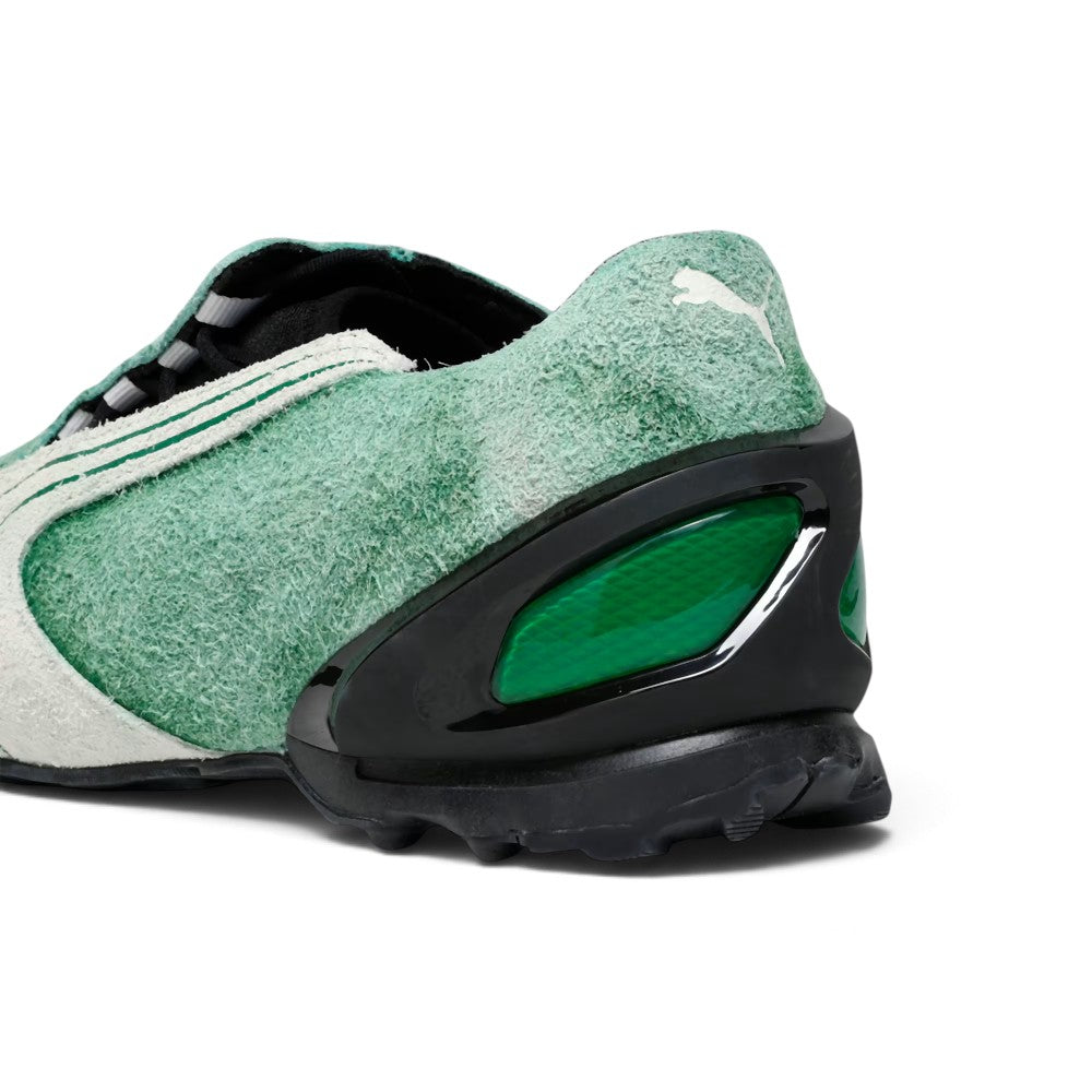 Close-up rear angle focusing on the green hairy suede upper, embroidered white PUMA Cat logo, glossy black TPU external heel counter with emerald-green cellular windows, and aggressively grooved black rubber heel tread.