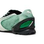 Close-up rear angle focusing on the green hairy suede upper, embroidered white PUMA Cat logo, glossy black TPU external heel counter with emerald-green cellular windows, and aggressively grooved black rubber heel tread.
