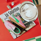 Dynamic editorial arrangement presenting the pair laid across an Italian sports newspaper beside a café tray, showcasing the bold pink suede chassis, emerald green leather Formstrip, fold-over flap tab, and printed green footbeds.