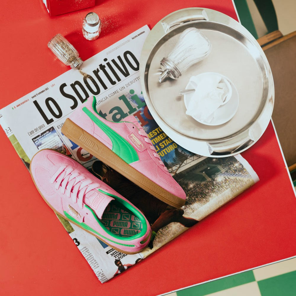 Dynamic editorial arrangement presenting the pair laid across an Italian sports newspaper beside a café tray, showcasing the bold pink suede chassis, emerald green leather Formstrip, fold-over flap tab, and printed green footbeds.