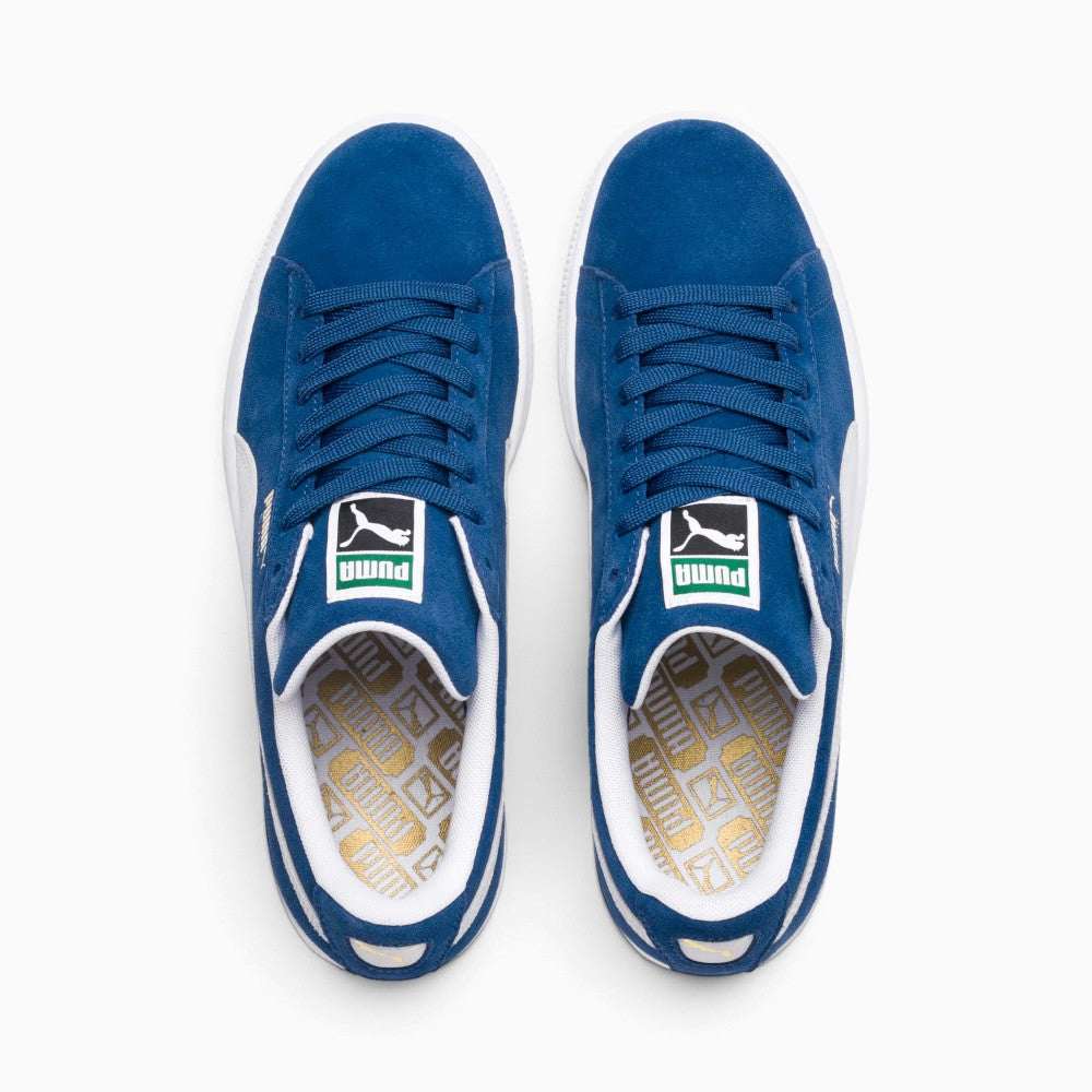 Direct overhead look at both shoes, displaying the Olympian Blue suede toe boxes, flat blue laces through punched suede eyestays, white-and-green woven PUMA tongue tags, and white footbeds stamped with an intricate gold foil repeating PUMA pattern.