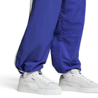 On-foot lifestyle angle showcasing both shoes paired with baggy blue track trousers, highlighting the exaggerated Y2K skate proportions, ultra-wide white fat laces, puffed foam tongue with green-and-white retro PUMA woven label, and bold low-top stance.