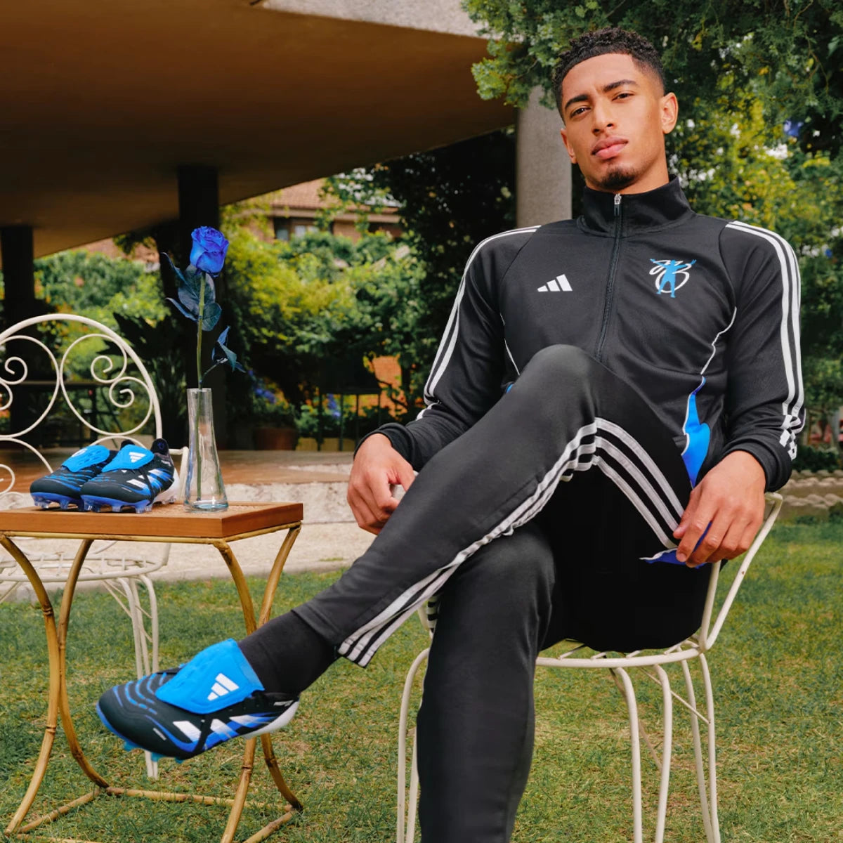 Lifestyle portrait of professional footballer Jude Bellingham seated outdoors, wearing a matching black-and-white track jacket and trousers while showcasing the signature Predator Elite boots on his feet alongside a blue rose.