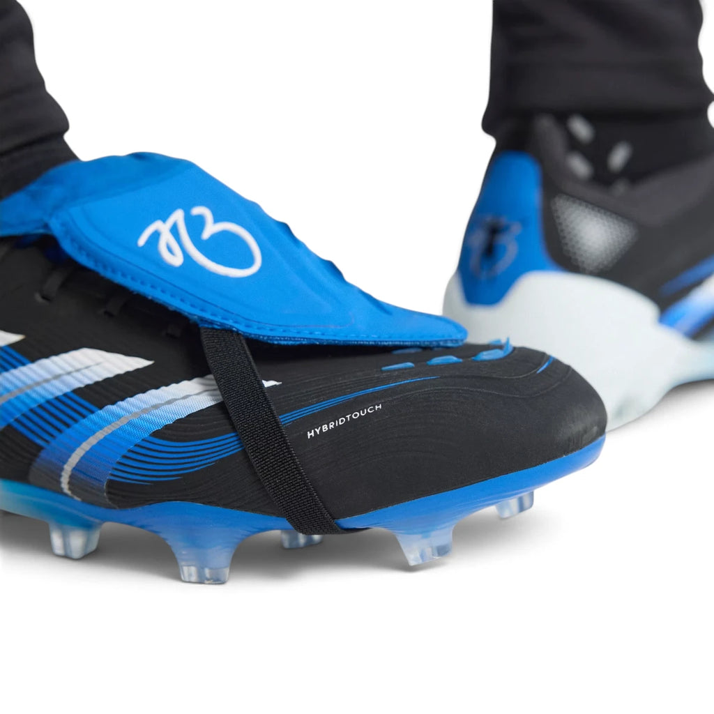 Close-up shot of a player's feet wearing the signature boots, focusing on the blue heel counter adorned with Jude Bellingham's iconic silhouette celebration graphic and the specialized firm ground studs.