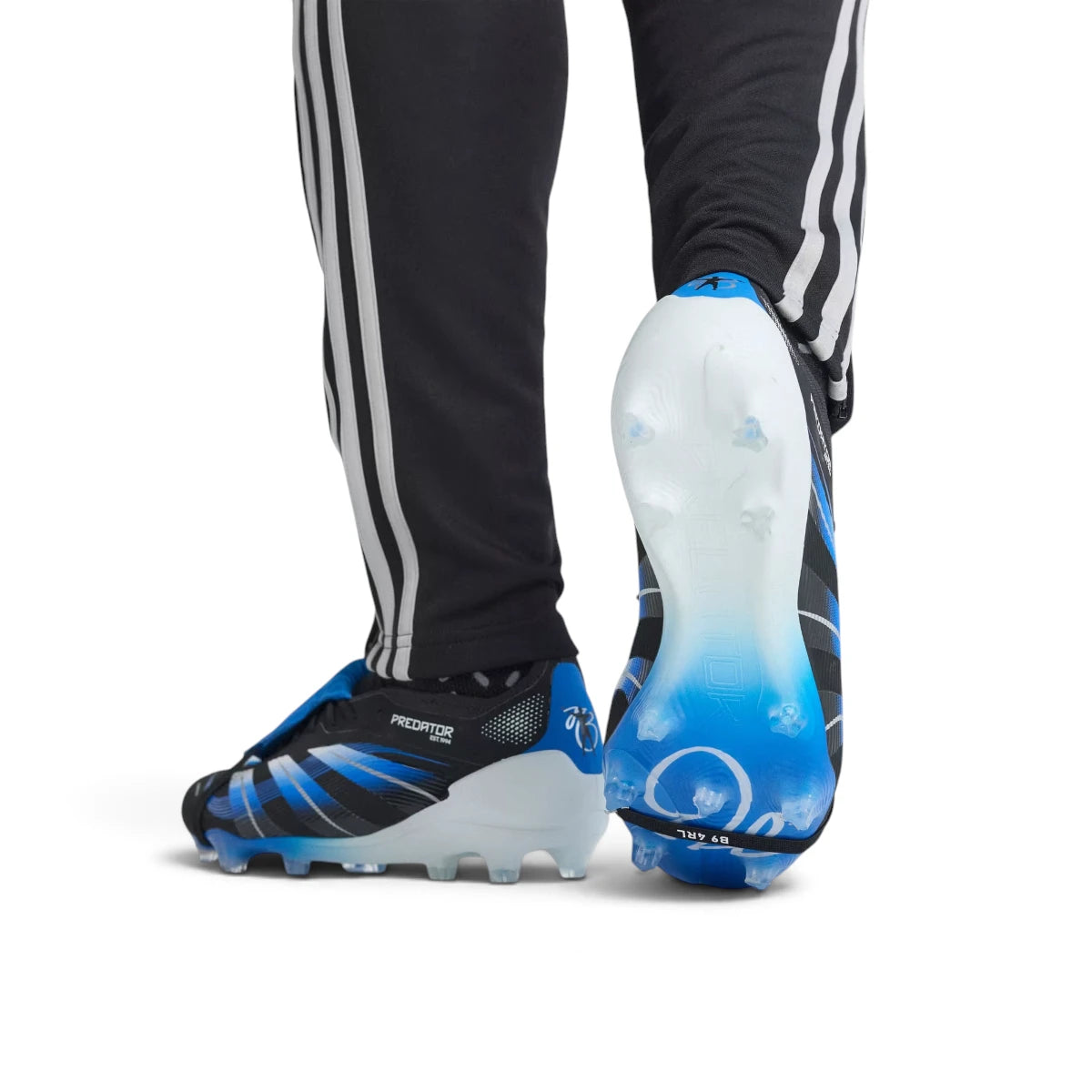Close-up shot of a player's feet wearing the signature boots, focusing on the blue heel counter adorned with Jude Bellingham's iconic silhouette celebration graphic and the specialized firm ground studs.