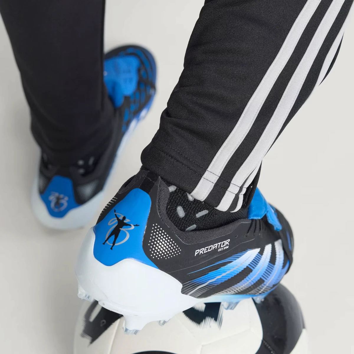Close-up shot of a player's feet wearing the signature boots, focusing on the blue heel counter adorned with Jude Bellingham's iconic silhouette celebration graphic and the specialized firm ground studs.