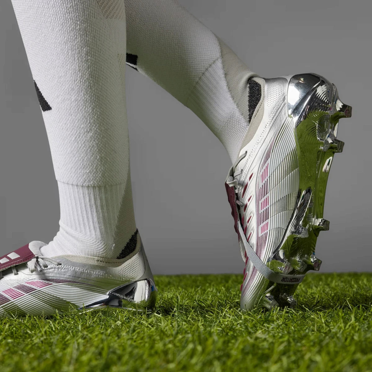 Close-up action perspective capturing a player running on grass, showcasing the reflective chrome outsole finish and silver upper profile.