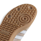 Close-up of the textured brown Adidas outsole showing the circular central tread and the embossed Adidas logo, with a white striped shoe upper partially visible underneath on a white background.