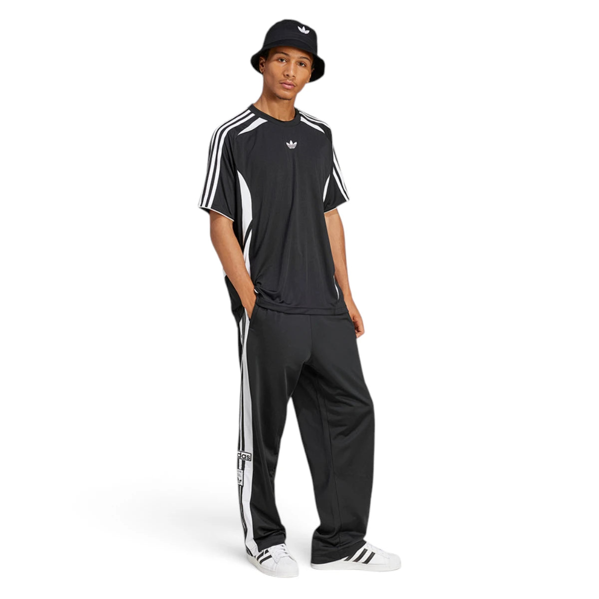 Full-length studio shot of a male model wearing the complete black Adibreak track look paired with a bucket hat and white shell-toe trainers, demonstrating its relaxed, everyday styling.