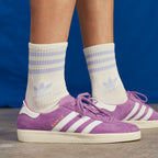 Purple Adidas Gazelle sneakers with white stripes, worn with white crew socks featuring light purple stripes and Adidas branding, set against a blue background.