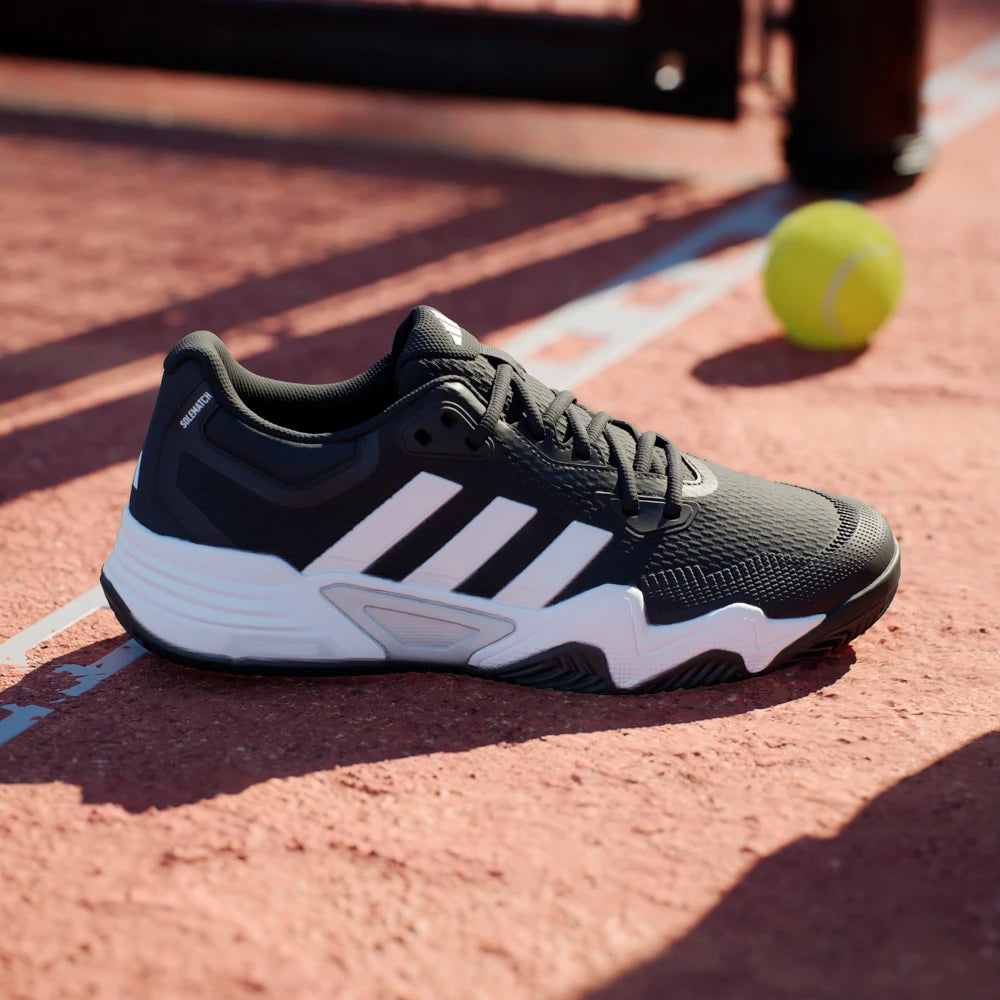 Black and white tennis shoe on a tennis court with a yellow ball in the background