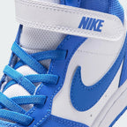 Close-up view of the forefoot, detailing the white leather toe box perforations, double-stitched Blue Crystal mudguard overlay, blue elastic laces, and white strap stamped with blue "NIKE" block lettering.