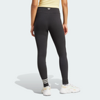 Rear view of the model wearing the black high-waisted adidas Neuclassics leggings, highlighting the small white adidas Trefoil logo printed on the back of the waistband and the asymmetric three white stripes around the left ankle.