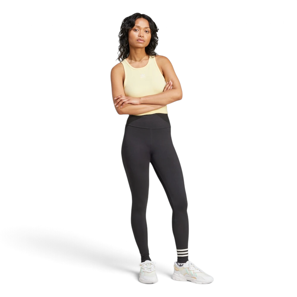 Full-length front view of the model standing with arms crossed, wearing a pastel yellow ribbed vest top, the black high-waisted adidas Neuclassics leggings, and chunky white trainers.