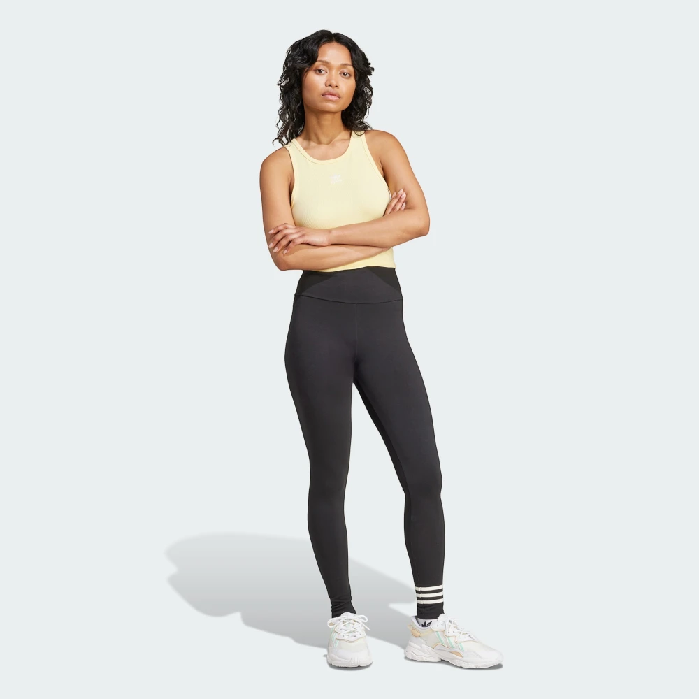 Full-length front view of the model standing with arms crossed, wearing a pastel yellow ribbed vest top, the black high-waisted adidas Neuclassics leggings, and chunky white trainers.