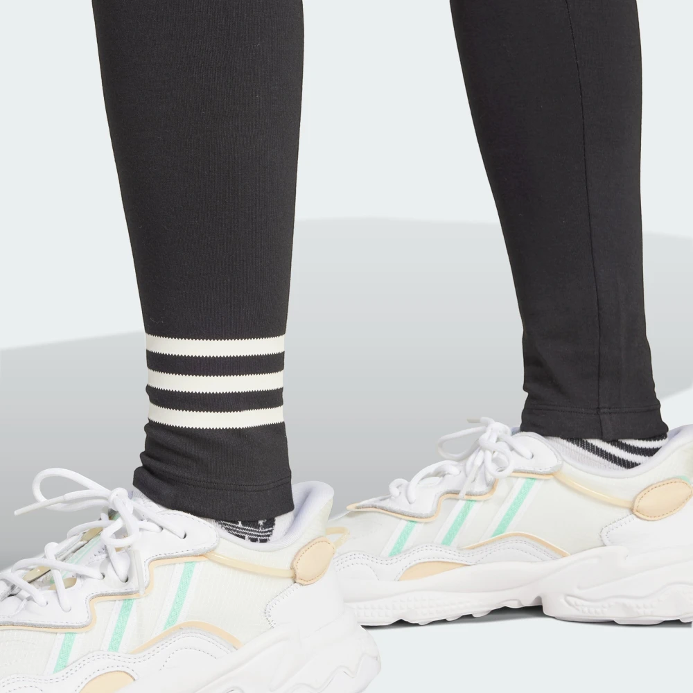Detailed close-up view of the lower legs and white trainers, highlighting the three contrasting white horizontal 3-Stripes wrapped around the left shin of the black leggings.