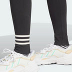 Detailed close-up view of the lower legs and white trainers, highlighting the three contrasting white horizontal 3-Stripes wrapped around the left shin of the black leggings.
