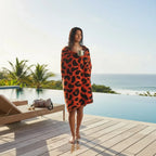 Woman wrapped in an orange towel with black patterns, standing by a pool with palm trees and ocean view.