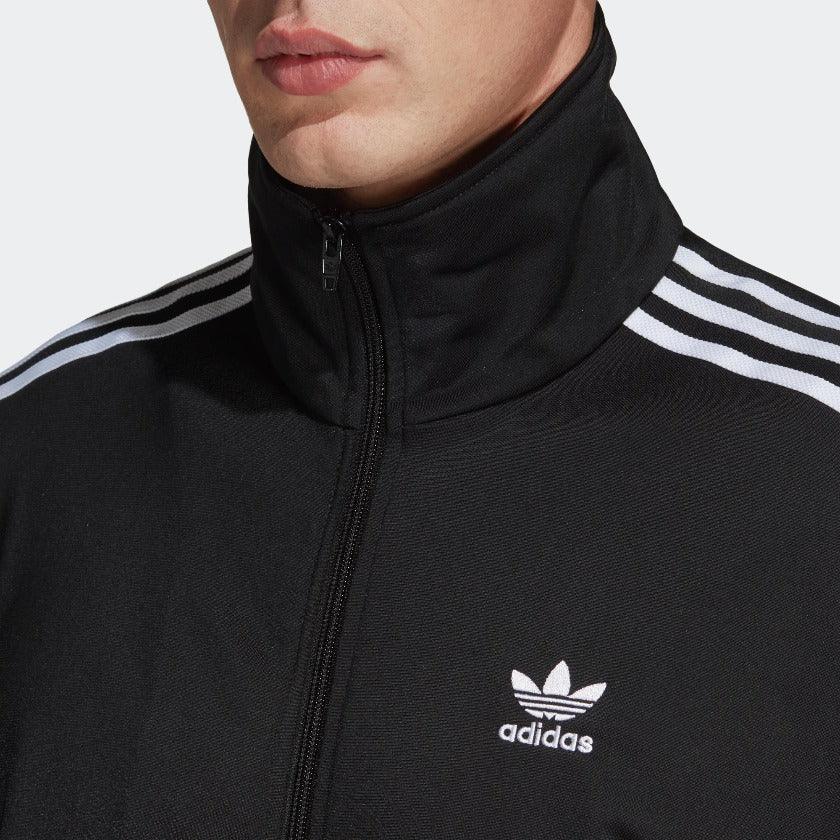 Close-up zoom on the fabric of the Adidas Firebird Track Jacket in black, showing the smooth tricot texture and the embroidered white Trefoil logo detail on the chest.