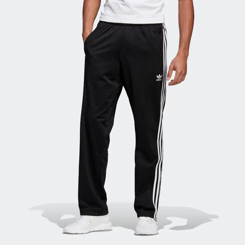 Adidas Firebird Track Pants Men's in black, model worn front view showing white 3-Stripes down the legs, Trefoil logo on the hip, and zipped pockets. Main image